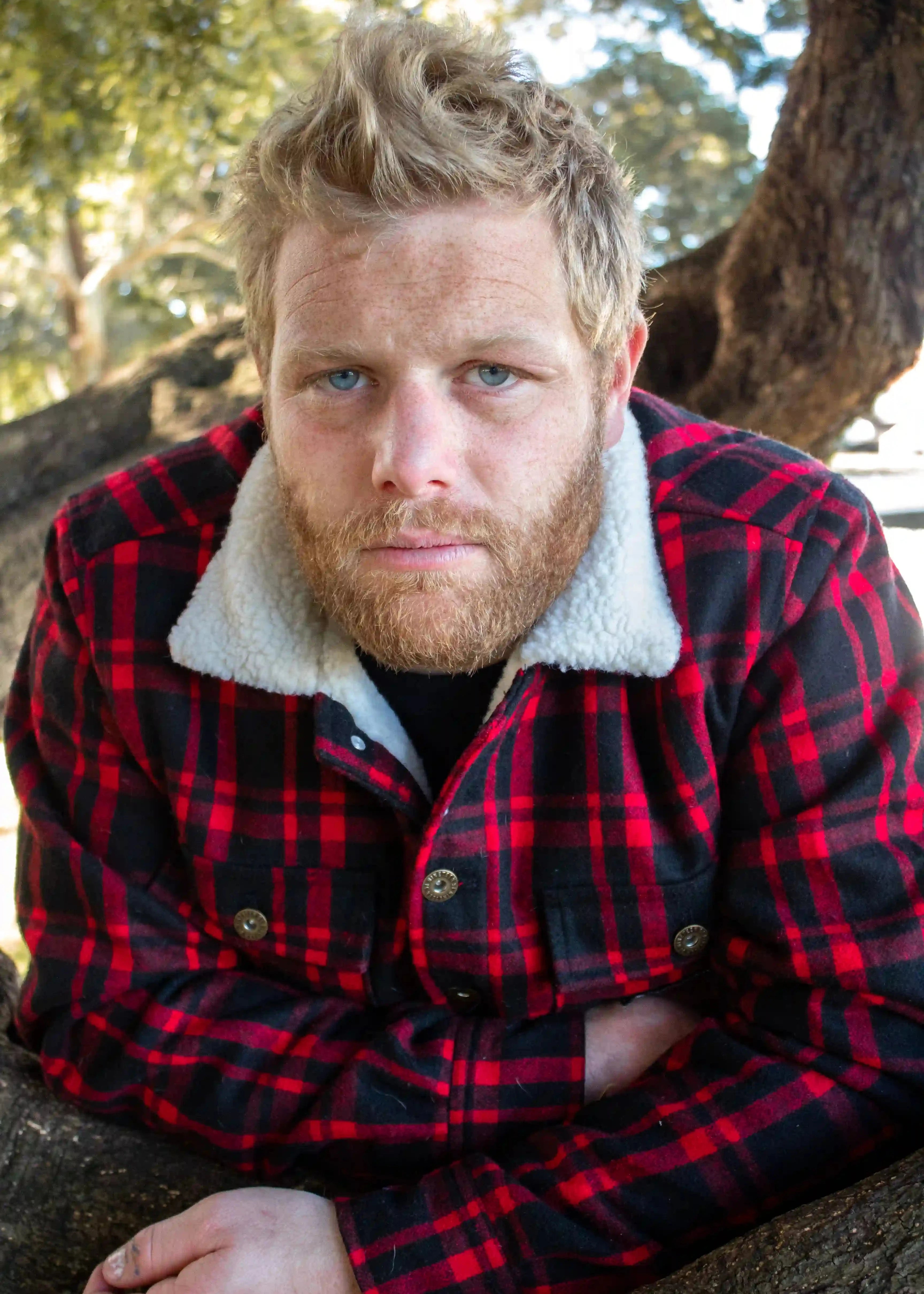 Man wearing a red and black plaid jacket with white fur lining, sitting outdoors.