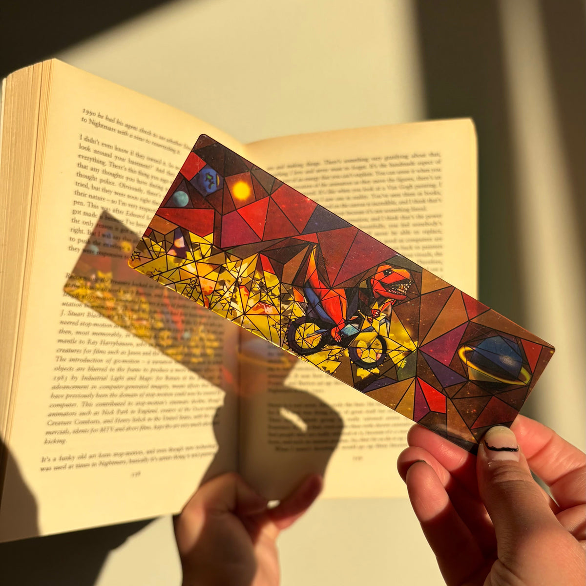 A person holds a colorful, geometric-patterned bookmark or art print in front of an open book, casting a shadow on the page. The design features a space theme with planets, stars, and a dinosaur in a stained-glass style.