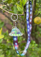 Green Dachshund flying a spcaeship acrylic keychain, attached to a long lanyard with a nebula background and paw prints walking up it.