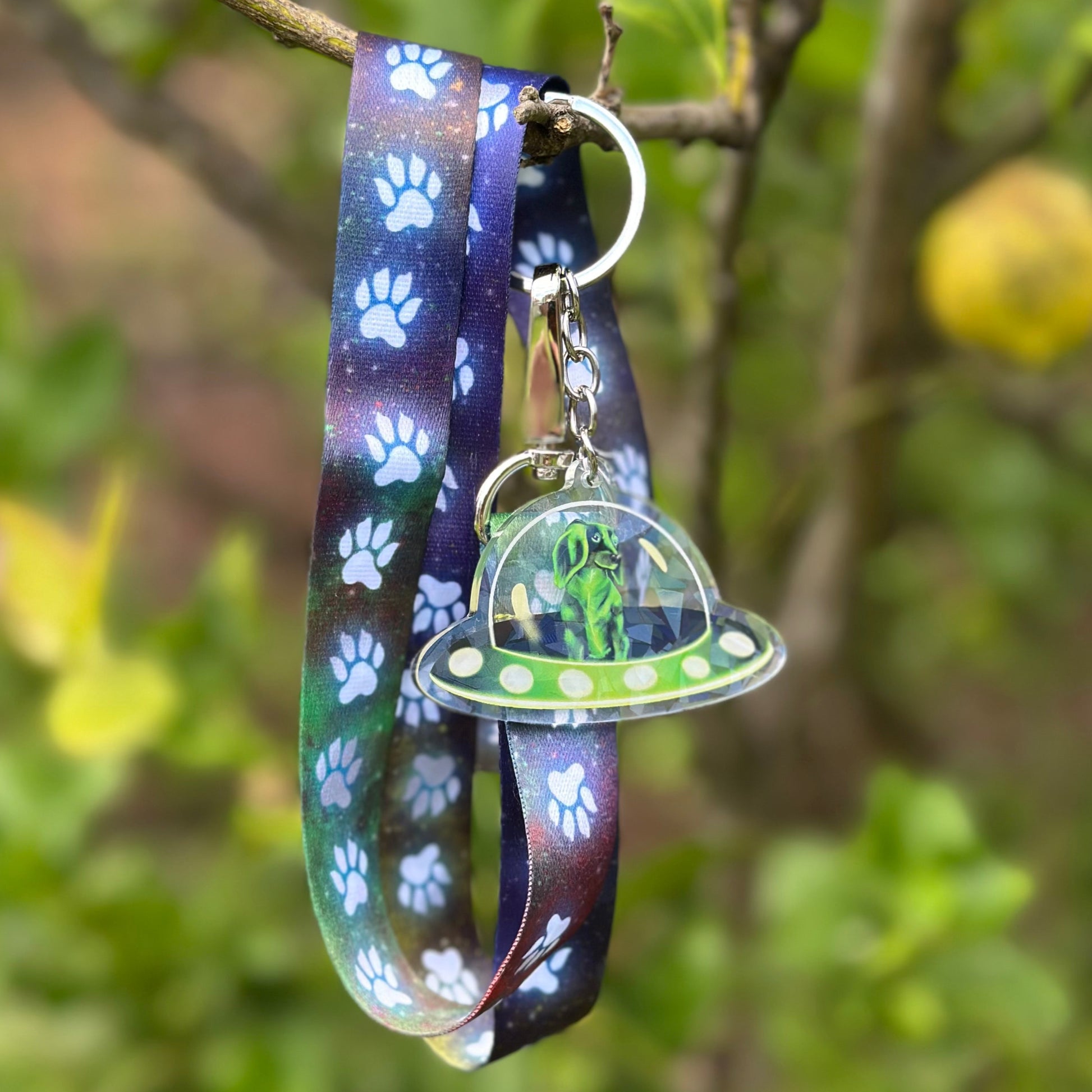 Green Dachshund flying a spcaeship acrylic keychain, attached to a long lanyard with a nebula background and paw prints walking up it.